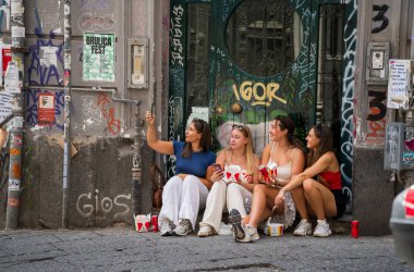Napoli, İtalya - 22 Haziran 2024: Dört kadından oluşan neşeli bir grup bir şehir sokağında oturup fast food paylaşıp fotoğraf çekiyorlar. Sahne, arkadaşlığı ve sokak kültürünü duvar yazısıyla süslenmiş bir ortamda yakalıyor..