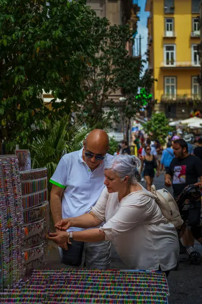 Napoli, İtalya - 22 Haziran 2024: Yaşlıca bir çift, canlı bir şehir ortamında açık bir sokak pazarında canlı mücevherlere göz atıyor, parlak mavi gökyüzünün altında sakin bir alışveriş yapıyor..