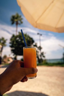 Bir bardak taze portakal suyunu kamışla tutan bir el. Parlak bir yaz günü, başımda bir şemsiye ve arkamda bulanık bir yeşillikle.
