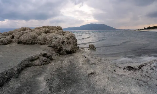 stock image Lake Salda, Turkey: A large rock formation leads into a body of water with a mountain in the distance under a cloudy sky, creating a tranquil and scenic coastal landscape.