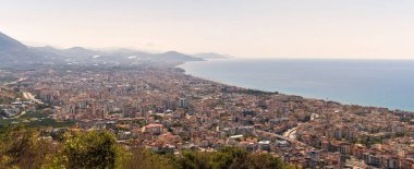 Türkiye 'nin Antalya kentinin panoramik hava görüntüsü, arka planda puslu bir gökyüzünün altında dağlar bulunan Akdeniz kıyı şeridi boyunca yoğun bir şehir manzarası sergiliyor.