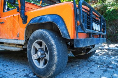 Engebeli turuncu 4x4 'lük aracın detaylı bir yakın çekimi off-road modifikasyonları, ön tampon ve lastiği vurguluyor, doğal bir şekilde taştan bir kaldırıma park edilmiş..