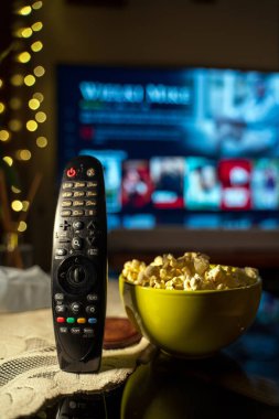 A close-up of a TV remote control on a table with a bowl of popcorn in the foreground, with a TV in the background showing a streaming service interface. clipart