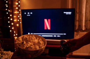 Krakow, Polonya - 15 Ekim 2024: Ön planda patlamış mısır ve arka planda Netflix logosunu gösteren bir televizyon kumandası tutan bir el.