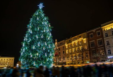 Büyük, ışıl ışıl dekore edilmiş bir Noel ağacı hareketli bir şehir meydanında durur, neşeli bir kalabalığın arasında güzel bir şekilde parlar ve büyüleyici bir tatil atmosferinde aydınlatılmış tarihi mimari ile çevrilidir..