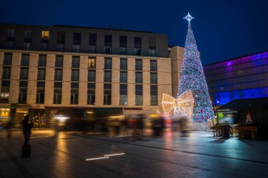 Şehir meydanını aydınlatan ışık ve yayla süslenmiş güzel dekore edilmiş bir Noel ağacı. Kuleler ve yakınlardaki binalar şenlikli bir şehir tatili ortamı yaratıyor..