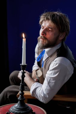 Dönem kıyafetleri içinde yansıtıcı bir adam, yumuşak bir ışık ve sakin bir atmosferle çevrili yıpranmış bir kitabı tutarak, düşünceli bir şekilde bir muma bakar..