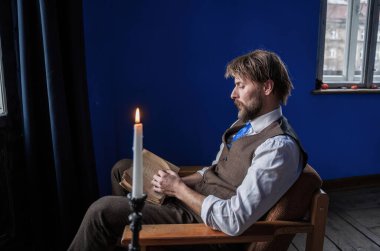 Kırsal giyinmiş düşünceli bir adam içeride oturur, mum ışığında eski bir kitap okur, mavi duvarlar ve pencerelerle çerçevelenir, sessiz ve içsel bir ruh hali uyandırır..