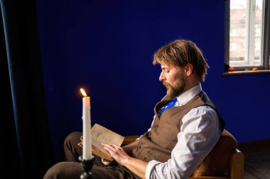 Kaliteli giyinmiş bir adam, mum ışığıyla aydınlatılmış koltukta eski bir kitap okur. Yumuşak doğal ışık sağlayan canlı mavi duvarlara yaslanır..