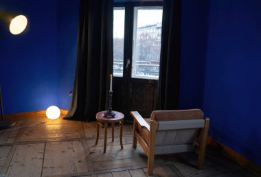 A cozy reading nook with a vintage armchair, a small table holding a lit candle, and a floor lamp casting a warm glow against a deep blue wall. clipart