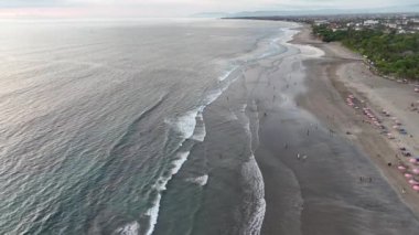 Bali 'deki Seminyak Sahili' nin üzerinde gün batımı altın gökyüzü ve parıldayan dalgalarla büyüleyici bir sahne yaratır. Sahile gidenler deniz kenarında mükemmel bir tropikal akşamın tadını çıkararak kıyı boyunca yürürler..