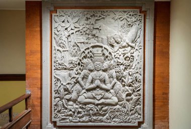 Geleneksel Bali tarzında üç meditasyon figürünü tasvir eden taş oyma. Karmaşık çiçek desenleri ve palmiye ağaçları ile çevrili. Ruhani sanatı yansıtıyor.