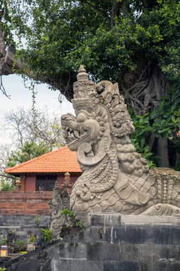 An ornate stone statue of a mythical beast in Bali, featuring elaborate designs and surrounded by traditional Balinese structures, including a red-roofed building and vibrant plants. clipart