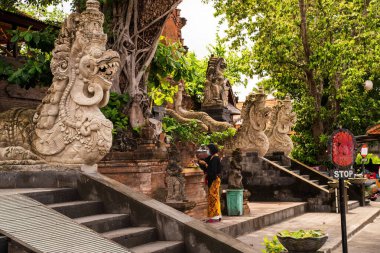 Bali, Endonezya - 29 Kasım 2023: Geleneksel Bali tapınağı gür yeşillikler arasında karmaşık ejderha heykelleriyle süslenmiş, bir kadın antik bir sunakta ruhani bir adak sunuyor..