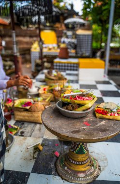 Balinese offerings on a rustic table, featuring vibrant food and woven crafts, await a ceremony. The checkered fabric and blurred figures in traditional dress add to the cultural scene in Bali. clipart