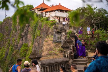 Bali, Indonesia - November 30, 2023: Bustling temple scene with vibrant-roofed structures atop dramatic cliffs, where visitors in traditional sarongs navigate historic steps while monkeys roam freely clipart