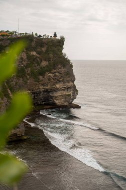 Uluwatu Temple perches dramatically atop a sheer limestone cliff, where crashing waves meet ancient rock formations while traditional Balinese architecture crowns the rugged coastline clipart