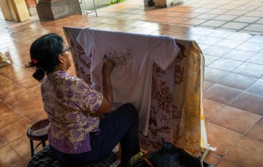 Bali, Endonezya - 23 Kasım 2023: Geleneksel Bali Batik 'i yaratan yetenekli zanaatkar, ahşap bir çerçeve boyunca uzanan beyaz kumaşa dikkatlice çiçek desenleri çiziyor.