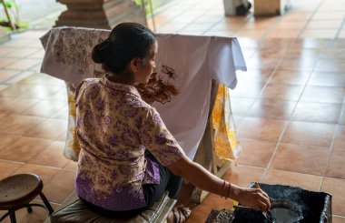 Bali, Endonezya - 23 Kasım 2023: Kalıplı bluz giyen kadın geleneksel batik sanatı için beyaz kumaş üzerine titizlikle çiçek hatları çiziyor.