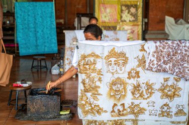 Bali, Endonezya - 23 Kasım 2023: Bir zanaatkar batik kumaş üzerinde çalışır, sıcak balmumu kullanarak efsanevi yüz, çiçek desenleri ve manzaralı manzaralar, tekstil sanatları sergiler..