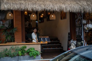 Bali, Endonezya, 11 Kasım 2023 sazdan çatılı tropik restoran girişi, bambu kolye ışıkları ve ahşap tezgah. Bir personel, saksı bitkilerinin menüsünün arkasında duruyor