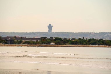 Bali 'nin ikonik Garuda Wisnu Kencana heykeliyle uzak tepenin panoramik manzarası. Kumlu sahilde hafif dalgalar dolaşırken tropikal tatil beldeleri ve sahildeki yemyeşil bitki örtüsü