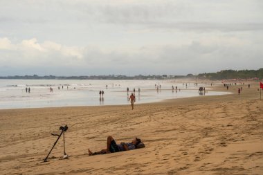 Bali, Endonezya, 11 Kasım 2023: Bali 'de sakin bir sahil sahnesi, dinlenen ve eğlenen insanların yer aldığı. Bulutlu gökyüzü sakin bir hava yaratırken bir kişi tripodun yanındaki kumda uzanır..