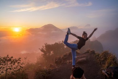 Bali, Endonezya, 11 Kasım 2024: Bir kadın, bulutlar ve sisle kaplı bir Bali dağının zirvesinde gün doğumunda yoga pozu veriyor. Altın saat ışığı huzurlu atmosferi güçlendirir, doğal güzelliği ve huzur ortamını ön plana çıkarır..
