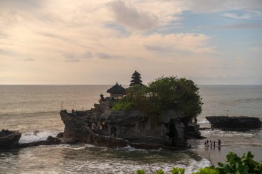 Dramatik Tanah Lot tapınağı engebeli taş kaidede okyanus dalgalarına karşı direniyor. Tropik yeşillik bu gizemli Bali deniz türbesini engin pastel gökyüzü altında çerçeveler..