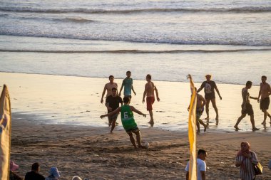 Bali, Endonezya - 11 Kasım 2023: Okyanus kıyısında sıradan bir futbol maçı: Oyuncular kıyıda dostça bir maçın keyfini çıkarırken diğerleri altın akşam ışığı altında seyrediyor.