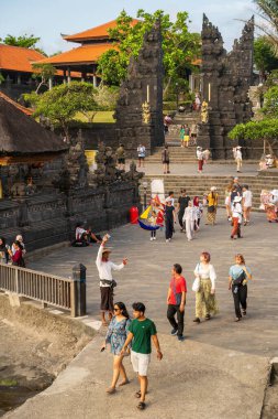 Bali, Endonezya - 11 Kasım 2023: Geleneksel bölünmüş kapıları ve turuncu çatılı pavyonları olan Antik Bali tapınağı girişi. Turistler taş avluyu keşfediyorlar.