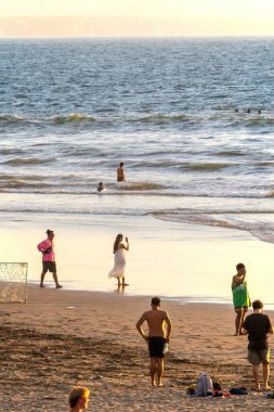 Bali, Endonezya - 11 Kasım 2023 Bali sahilinde gün batımının tadını çıkaranlarla altın saat. Beyazlı bir kadın fotoğraf çekerken diğerleri hafif dalgalarla yürüyor..