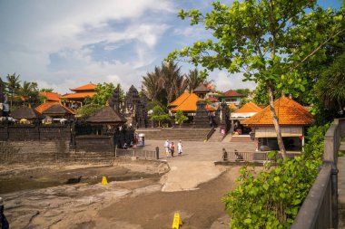 Bali, Endonezya - 11 Kasım 2023: Süslü taş kapıları olan Antik Bali deniz tapınağı ve mavi gökyüzü altında kutsal toprakları keşfeden beyaz elbiseli turistleri karşılayan geleneksel pagodalar