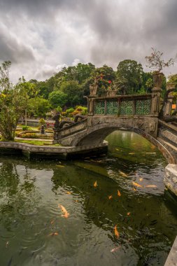 Bali 'de, oyma bir taş köprü koi dolu bir gölet boyunca uzanır, yoğun ağaçlarla dolu canlı bahçeler ve dramatik bulutlu bir zemin. Tirta Gangga Su Sarayı