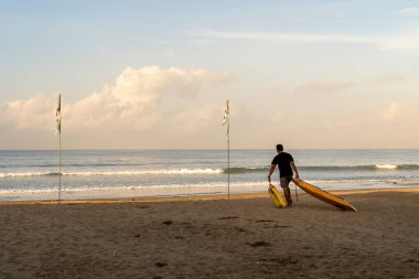 Bali, Endonezya - 12 Aralık 2023: Bir adam Bali 'de gün doğumunda sörf tahtasını iki bayrak arasındaki nazik dalgalara doğru sürüklüyor..