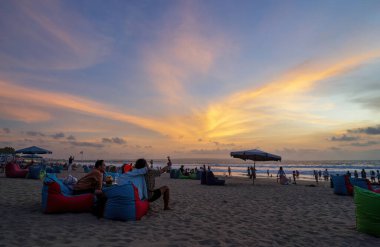 Bali, Endonezya - 12 Aralık 2023: Rahat puf minderler üzerindeki turistler, altın ışığın gökyüzünü boyadığı ve yumuşak dalgaların kumlu kıyı şeridini kapladığı muhteşem Bali günbatımının tadını çıkarıyorlar.