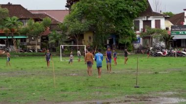 Bali, Endonezya - 12 Aralık 2023: Yerel çocuklar geleneksel Bali evleri ve tapınak mimarisi zeminiyle yeşil köy futbol maçının keyfini çıkarıyorlar