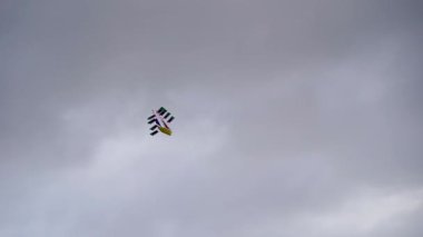 Bulutlu bir gökyüzüne doğru yükselen yelkenli gemi şeklinde canlı renkli bir uçurtma. Sahne, dramatik bulutlu havayla çelişen tuhaf bir atmosfer yakalıyor..