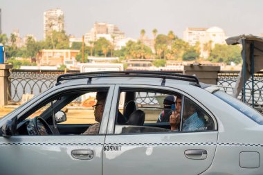 Kahire, Mısır - 11 Eylül 2023: Nehir kenarında kalabalık bir caddeye park etmiş yolcularla dolu şehir taksisi, yeşillik, kentsel mimari ve canlı yaşamla çevrili, benzersiz bir anı yakalıyor.