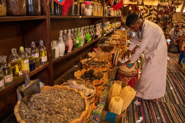 Kahire, Mısır - 11 Eylül 2023: Mısır pazarında çeşitli kurutulmuş bitkiler, baharatlar, yağlar ve el yapımı eşyalar sergilenmektedir. Geleneksel giyinmiş bir adam, eşyaları dikkatlice sergiliyor.