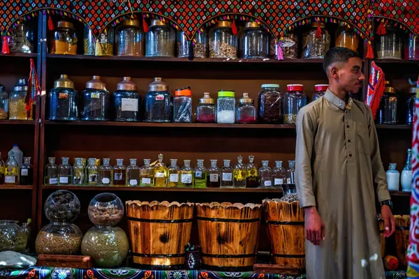 Kahire, Mısır - 11 Eylül 2023: Kokulu otlar ve baharatlarla dolu kavanozlar, canlı kültürel atmosferli ve geleneksel giysiler içinde duran bir tüccar ile karışık vitrinler.