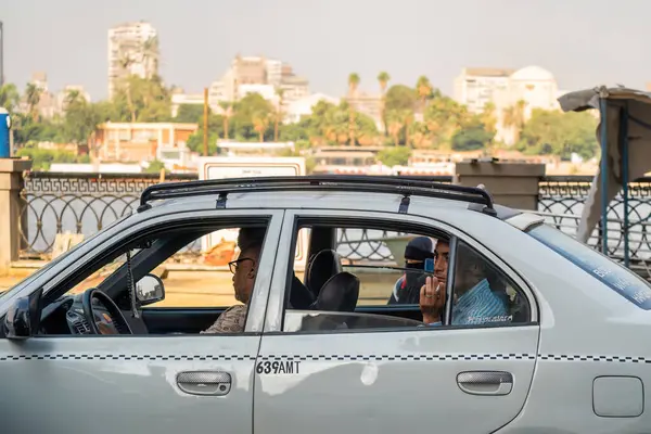 Kahire, Mısır - 11 Eylül 2023: Nehir kenarında kalabalık bir caddeye park etmiş yolcularla dolu şehir taksisi, yeşillik, kentsel mimari ve canlı yaşamla çevrili, benzersiz bir anı yakalıyor.
