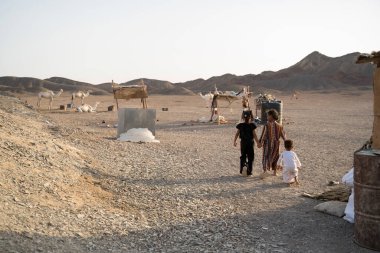 Kahire, Mısır - 11 Eylül 2023: Üç çocuk el ele yürür çölde, yakınlarda dinlenen develer ve derme çatma barınaklar ile birlikte, engebeli bir sadelik ve göçebe çöl hayatı hissi uyandırır..