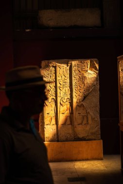 Kahire, Mısır - 9 Eylül 2023: Hiyeroglifler ve oyulmuş insan figürlerinin yer aldığı aydınlatılmış Mısır taş levhası, bir müzede sergileniyor, ön planda bir ziyaretçinin silüeti bulunuyor..