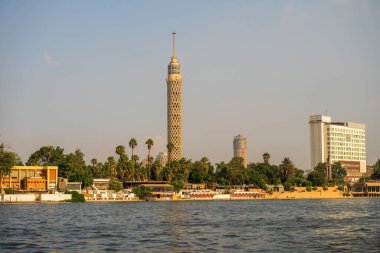 Kahire ufuk çizgisinde ikonik Kahire Kulesi, bereketli palmiye ağaçları ve Nil Nehri kıyısı berrak bir gökyüzü altında, Mısırın modern mimarisi ve doğal güzelliği sergilenmektedir.