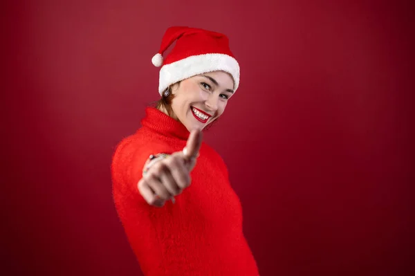 Young positive woman in christmas santa hat show thumb up like gesture isolated over red wall background. Happy young woman in Santa\'s helper hat a red background. People and winter holidays concept