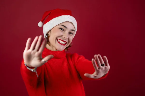 戴着圣诞圣塔帽的年轻而积极的女人被隔离在红墙的背景之外 快乐的年轻女人戴着圣诞老人的助手帽 背景是红色的 人与寒假概念 — 图库照片