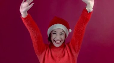 Noel Baba şapkalı genç pozitif kadın kırmızı duvar arkasında izole edilmiş. Noel Baba 'nın yardımcı şapkalı mutlu genç bayan kırmızı arka planda dans ediyor ve şımartıyor. İnsanlar ve kış tatili konsepti