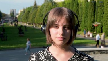 Dışarıda kulaklık takan genç bir kızın portresi. Kameraya bakmak ve modern kulaklıkla müzik dinlemek.