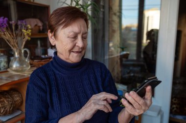 Akıllı telefon kullanan yaşlı bir kadının portresi. Pencerenin yanındaki yaşlı kadın cep telefonu kullanıyor.